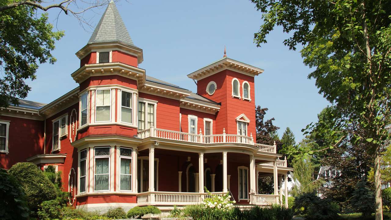 A large red victorian mansion with white shutters and a balcony and trees around