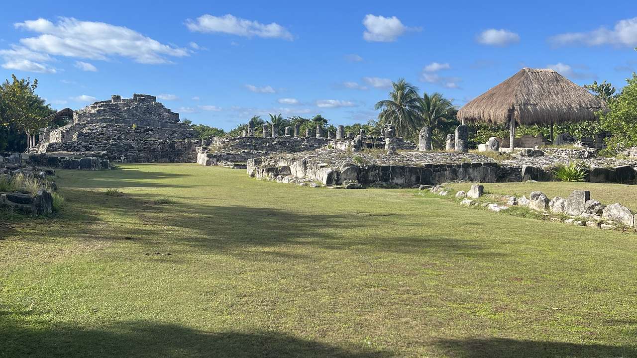 Side view of ancient ruins with trees and grass all around, and a hut to the right