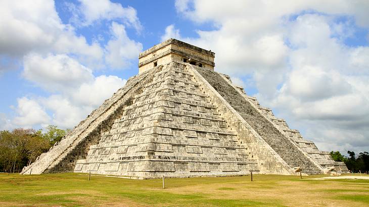 A Mayan pyramid structure with grass around it, under a blue sky with clouds