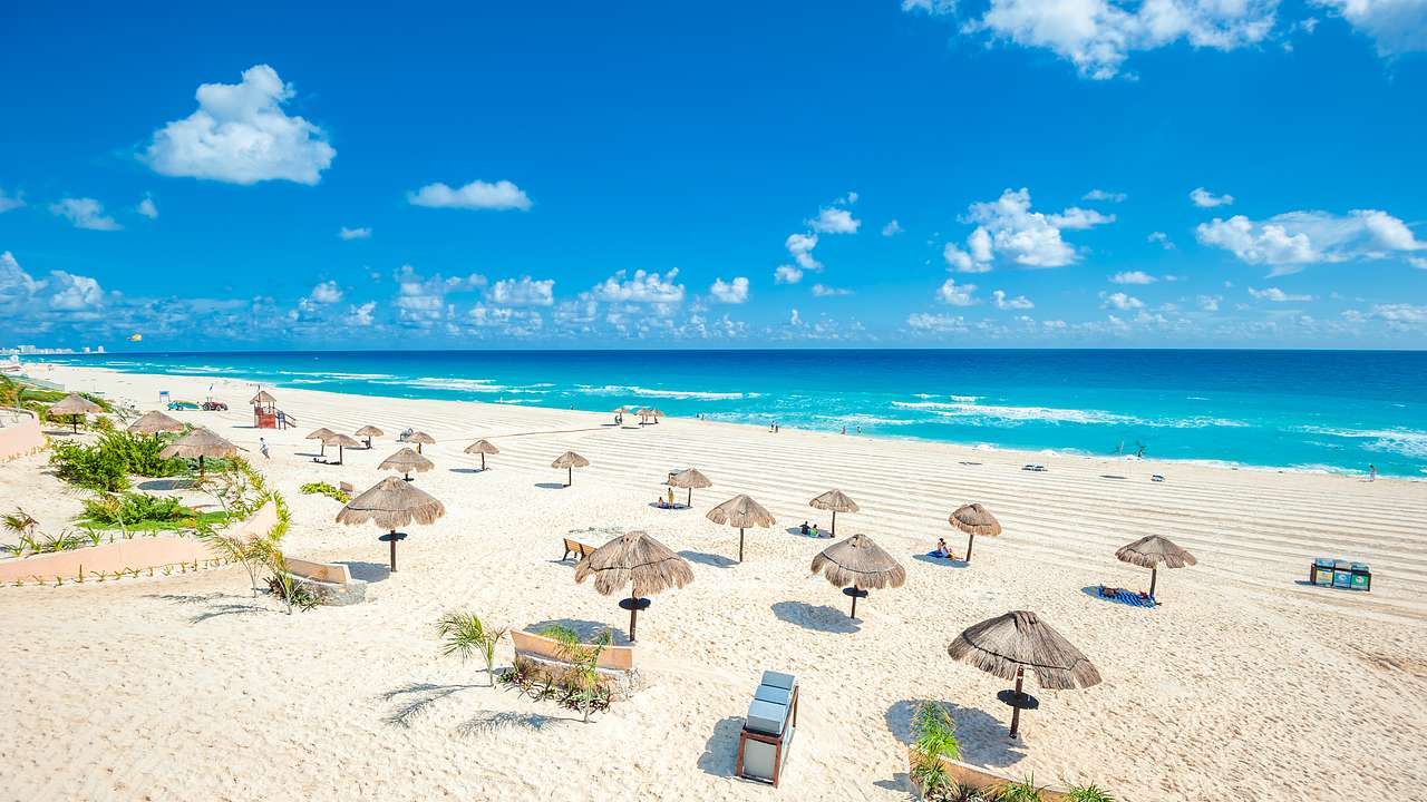 A beach with white sand, straw beach umbrellas, and blue ocean under a blue sky
