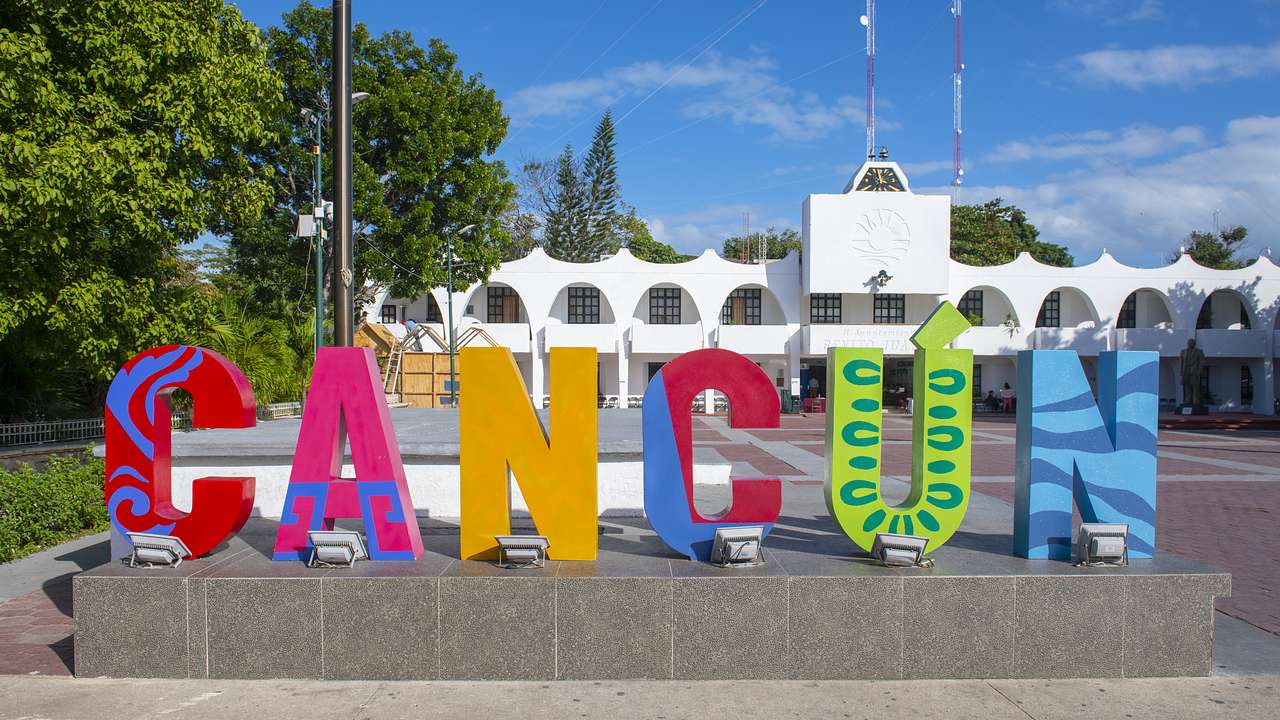 A 3D colorful Cancun sign with a white building and trees behind it