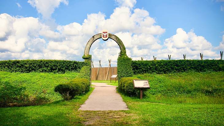 Green grass and bushes with an archway in the middle under a blue sky with clouds