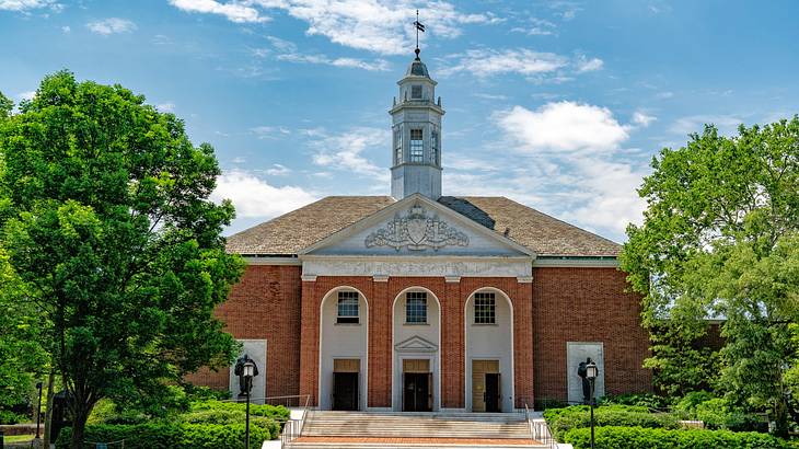 A redbrick building with columns and a white tower next to steps and green trees