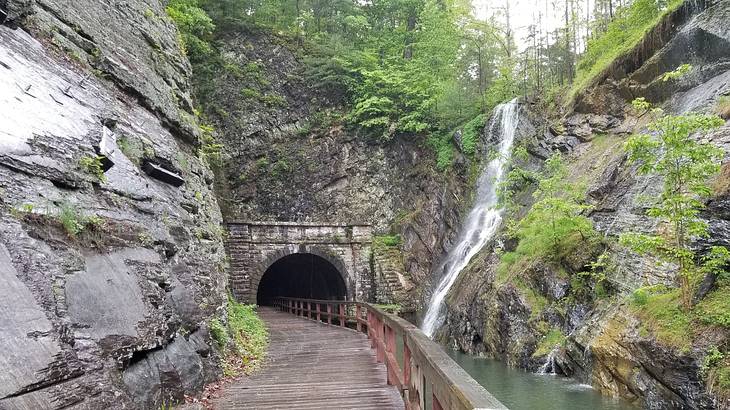 A wooden pathway next to a river and waterfall with a small tunnel at the end