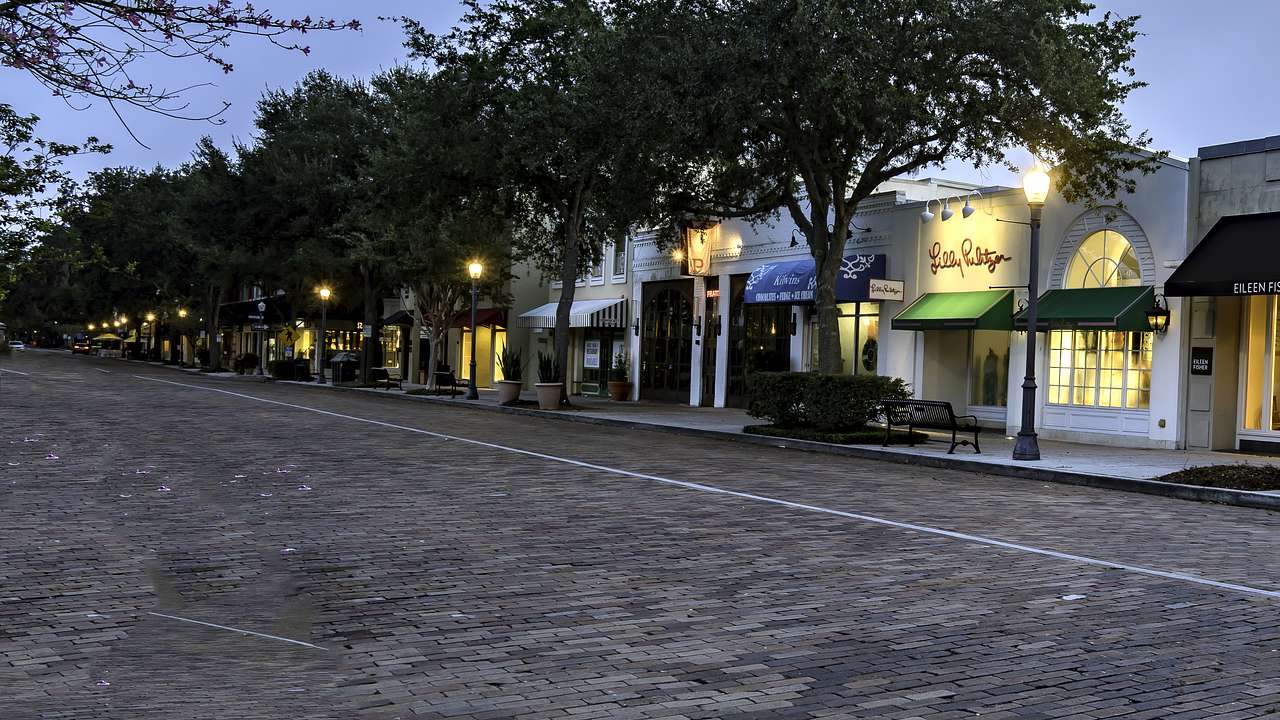 Boutique stores with illuminated street lamps along a bricked road lined by trees