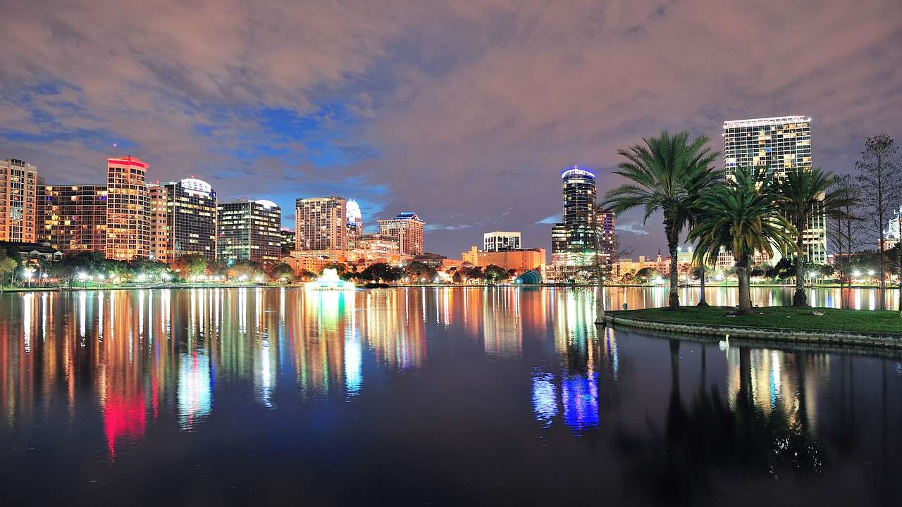 City buildings illuminated at night reflecting on a lake next to palm trees