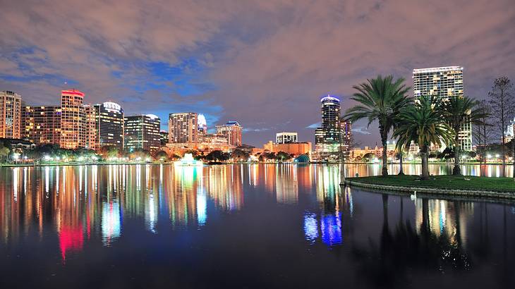 City buildings illuminated at night reflecting on a lake next to palm trees