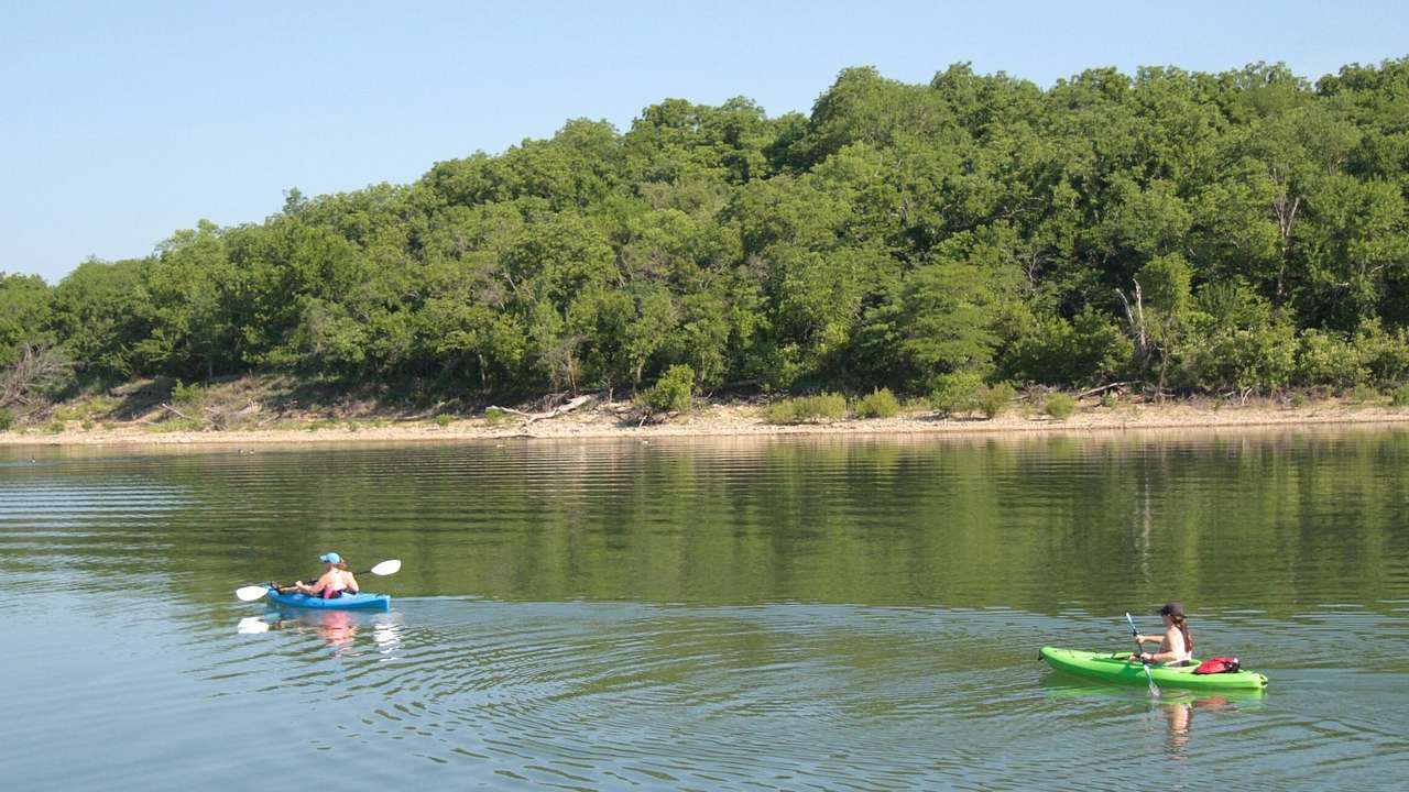 Two people kayaking on a lake with green trees behind on the shore