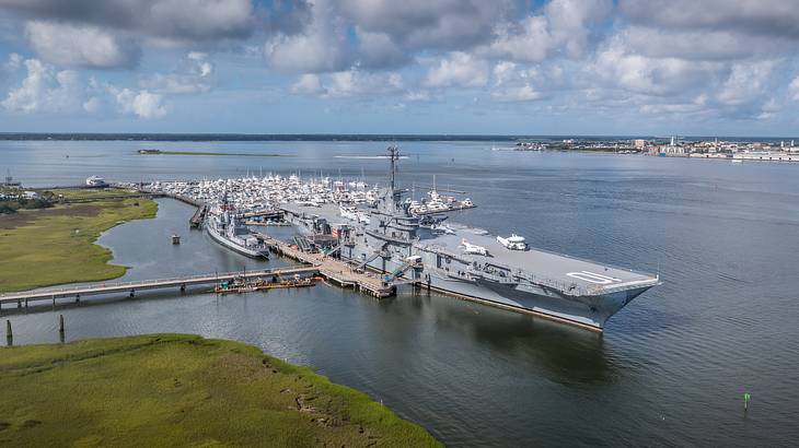Aerial of an aircraft carrier docked at a small pier under a partly cloudy sky
