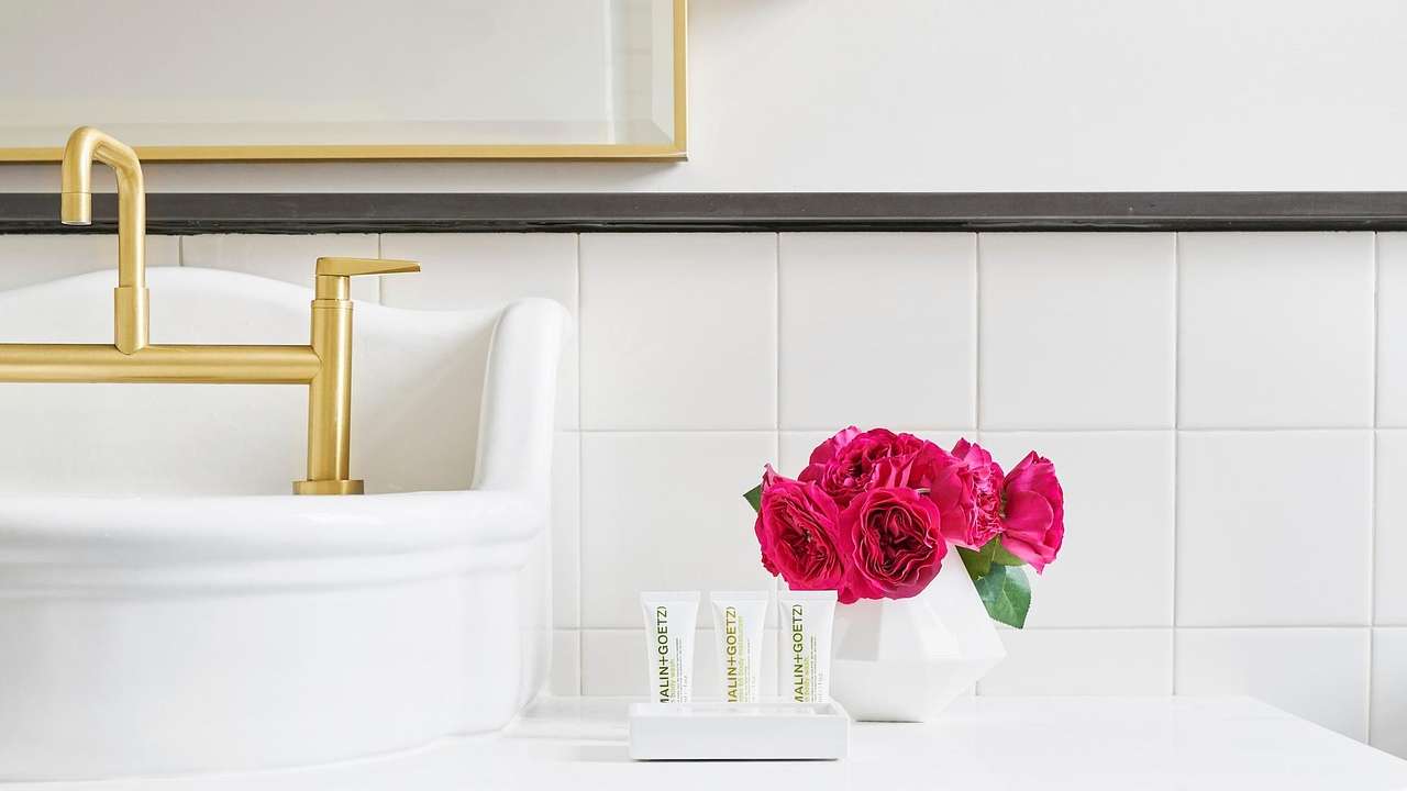 A bathroom with white tiles, a white sink and gold tap, and pink flowers