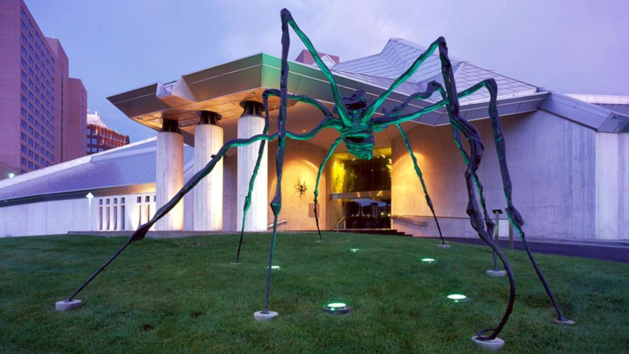 A spider sculpture in front of an illuminated museum at dusk
