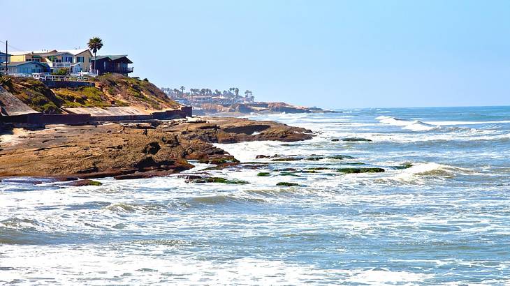 The ocean with waves next to a rocky cliff with a house and a palm tree on it