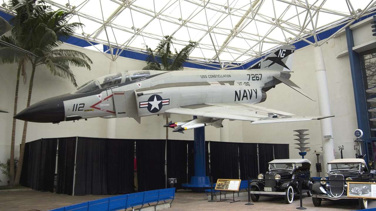 A Navy aircraft in a museum with trees and old-fashioned cars next to it