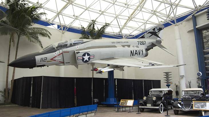 A Navy aircraft in a museum with trees and old-fashioned cars next to it