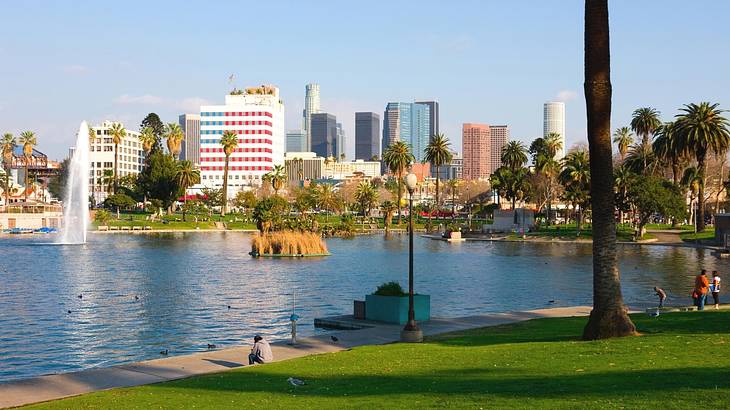 A park with a large pond with a fountain in it and city buildings behind it