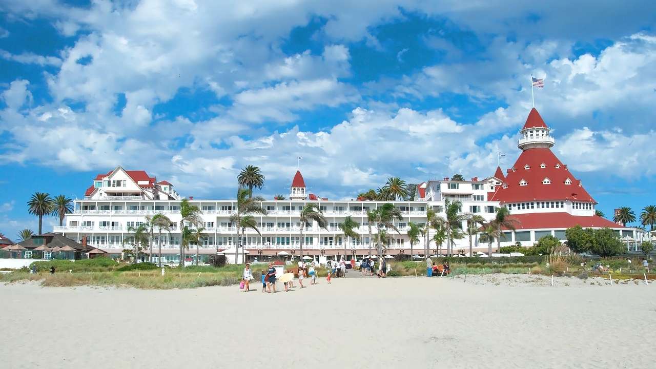 A white hotel building with a red roof next to a beach with palm trees