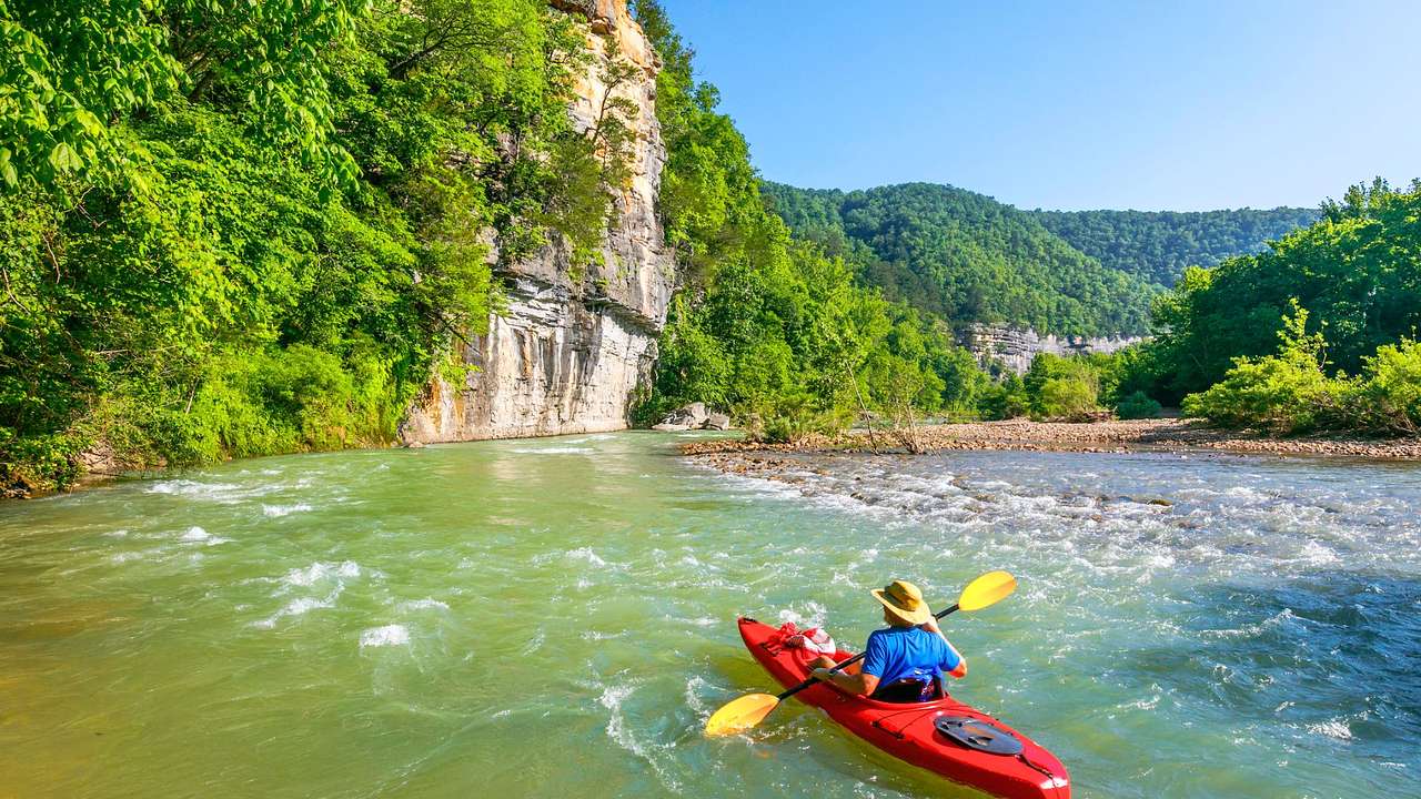 A person on the water in a red kayak surrounded by greenery-covered cliffs