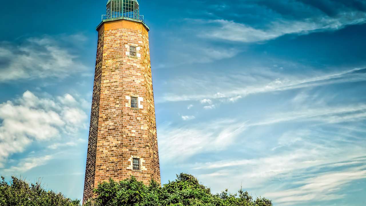 A tall, light-brown brick lighthouse with bushes in front under a clear blue sky