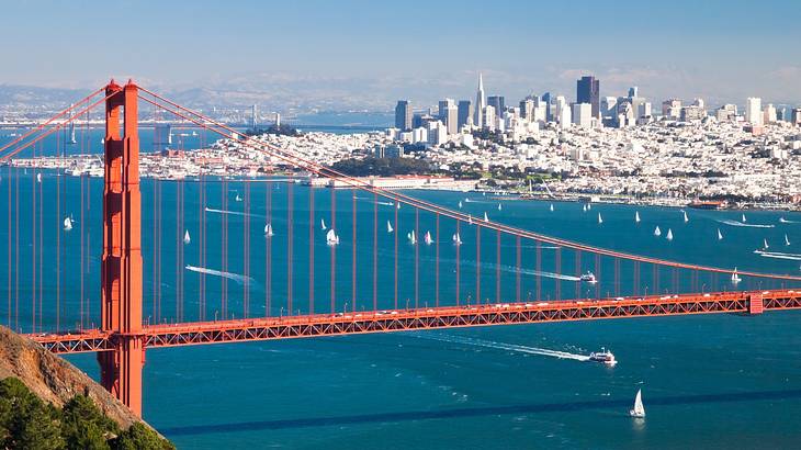 A red suspension bridge over the water with a city in the distance