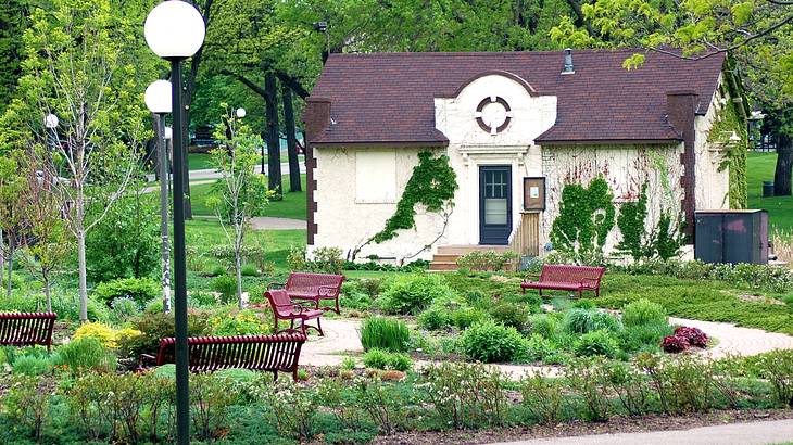 A white building with a brown roof surrounded by a lush green garden and lamp posts