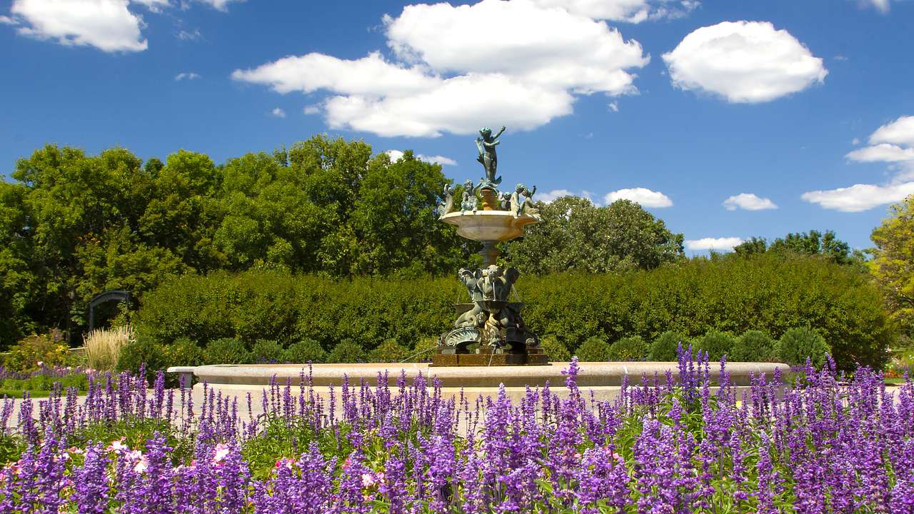 A fountain with sculptures in it surrounded by violet flowers and trees at the back