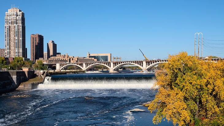 A small waterfall over a river next to trees and buildings under a blue sky