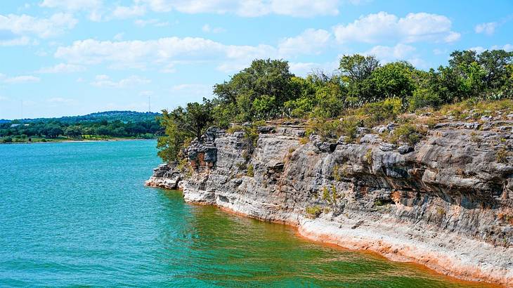 A lake with rocky cliffs on the right that are covered in trees under a blue sky