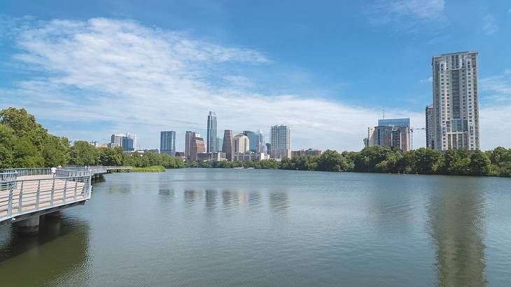 A lake with trees and a boardwalk to the side and a city skyline in the distance