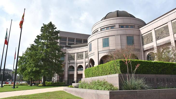 A museum building with a rotunda and trees, bushes, and flags in front of it