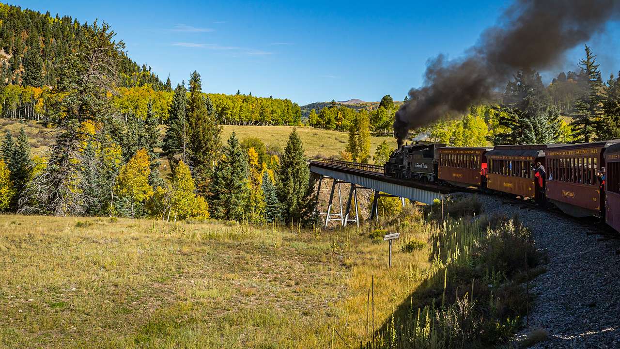 A steam train on a rail road winding through green hills with trees under a clear sky