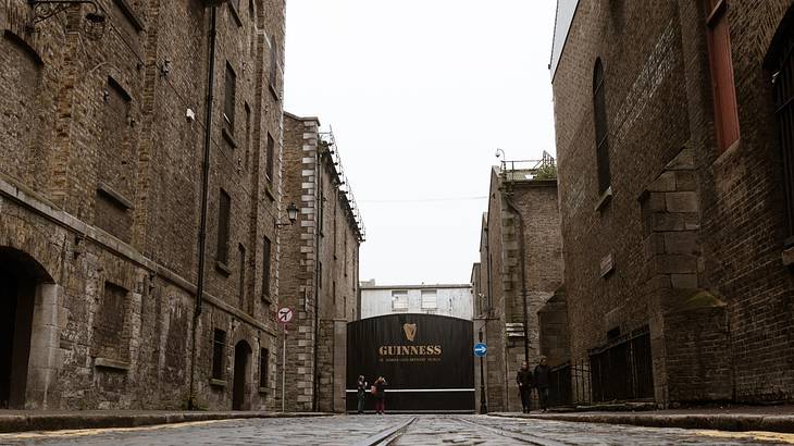 A brick road lined with buildings, leading to a gate with a Guinness sign from below