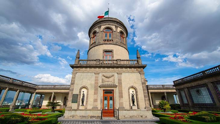 A neoclassical-style castle with the Mexico flag on top, in the middle of a lawn