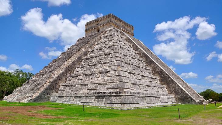 An ancient limestone pyramid structure surrounded by grass under a partly cloudy sky