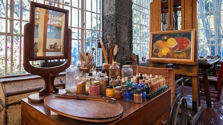 A table with painting utensils, a mirror, & a painting of fruits hanging on the right