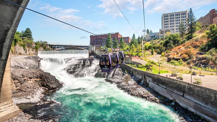 Cable cars over a waterfall and pool with greenery and buildings to the side