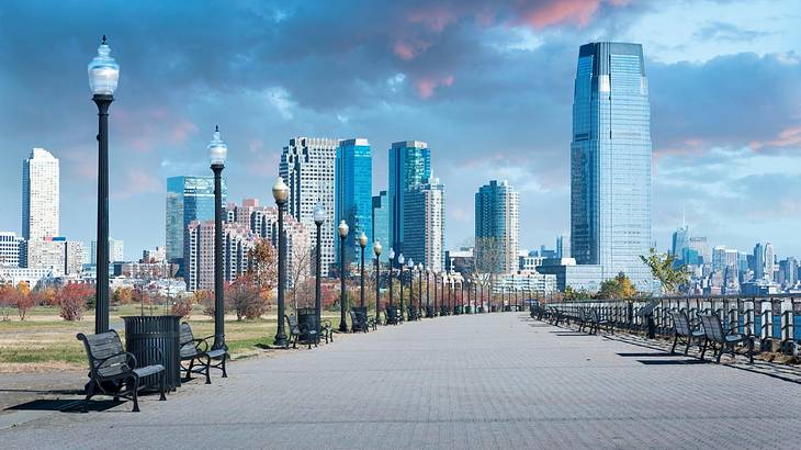 A path through a park lined with benches and lampposts and a skyline at the end
