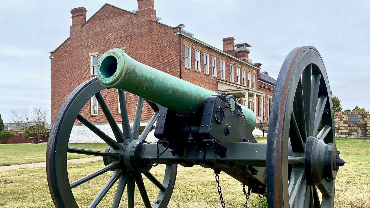 A black and green field cannon on a grassy lawn and a brick building in the back