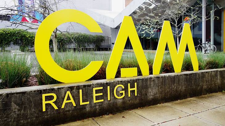 A large yellow sign that says "CAM Raleigh" on a wall next to grass and a path