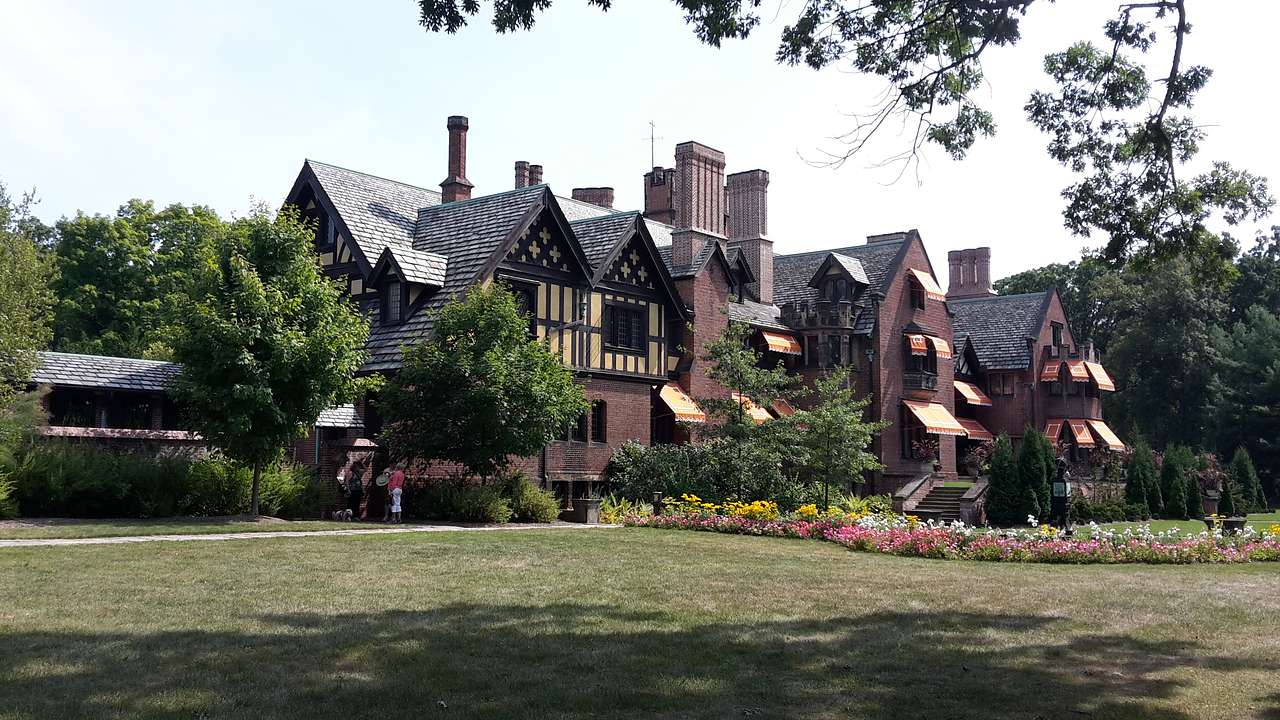 A brown mansion and carriage house with a row of colorful flowers and a green lawn