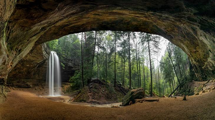 A huge sandstone cave opening facing trees and a small waterfall