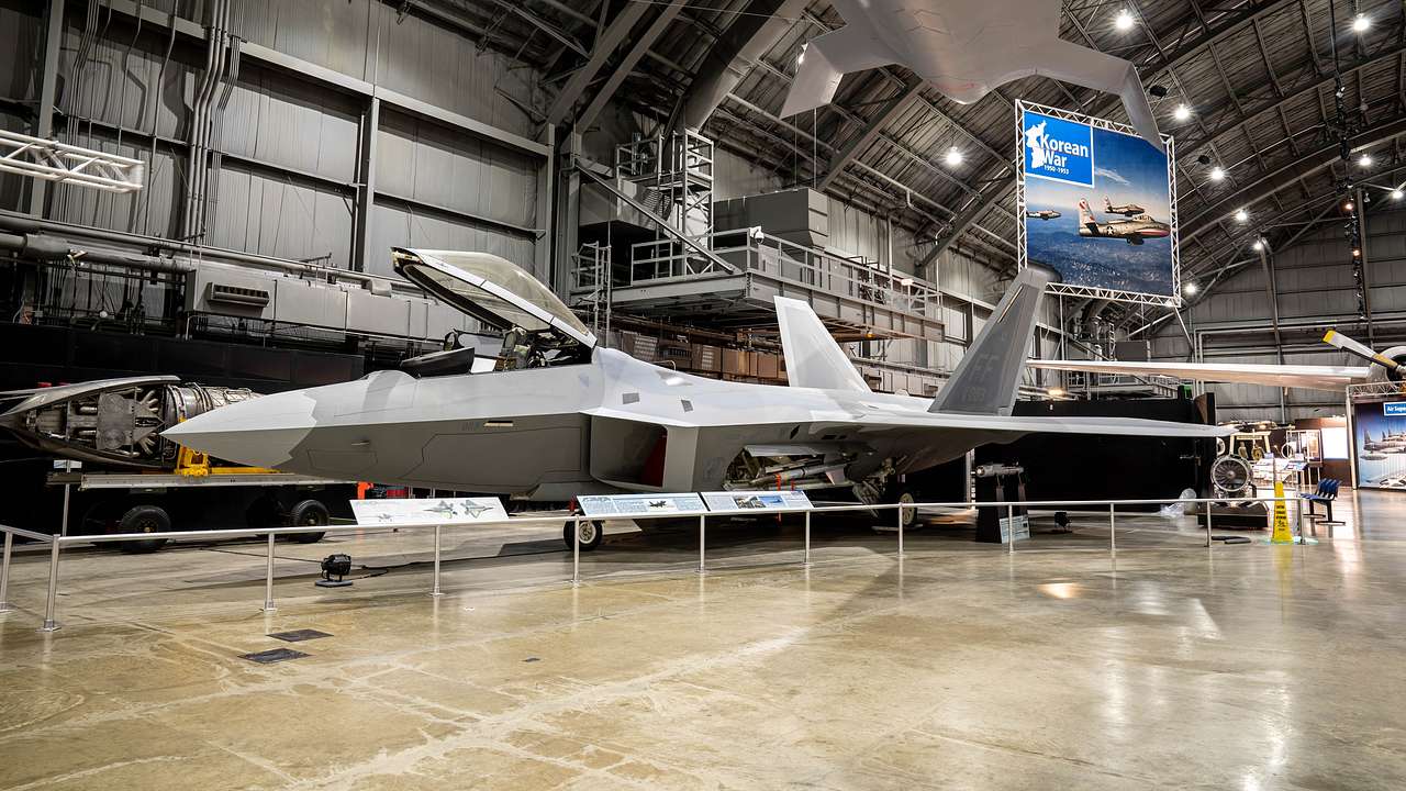 A hangar with a fighter aircraft on the ground and an aircraft hung above.