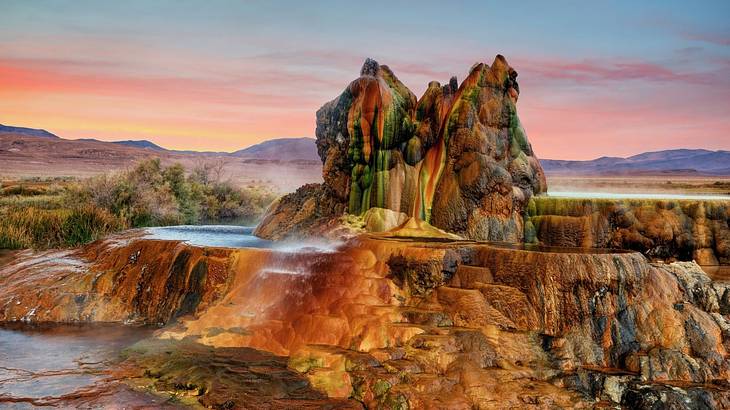 Red and green geyser mound with a steaming pool and mountains behind