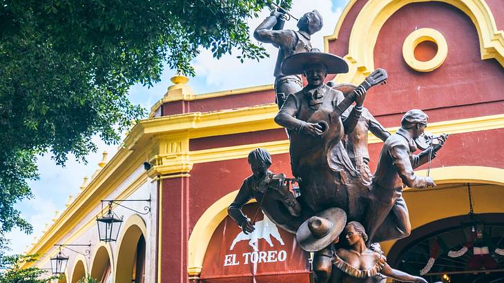 A bronze statue of a Mariachi band next to a tree and a red and yellow building