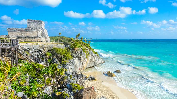 Ruins on top of a headland with greenery, near a white sand beach next to blue water