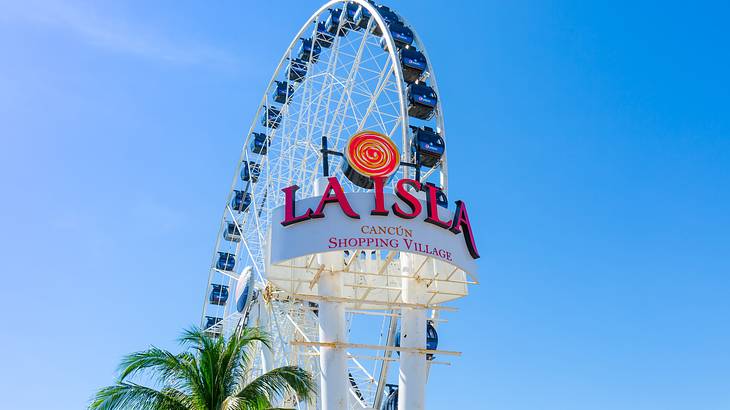 A sign that says "La Isla Shopping Village" with a Ferris wheel behind it