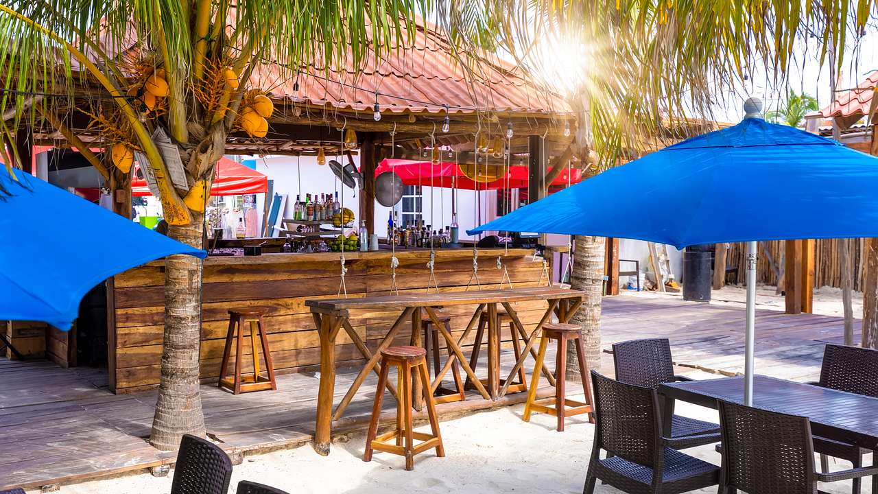 Brown chairs and tables under blue umbrellas, next to a tiki bar and trees