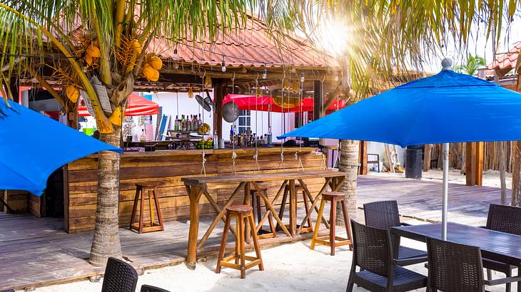 Brown chairs and tables under blue umbrellas, next to a tiki bar and trees