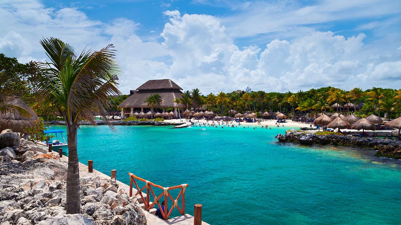 Turquoise water surrounded by sand, palm trees, and huts on the shore