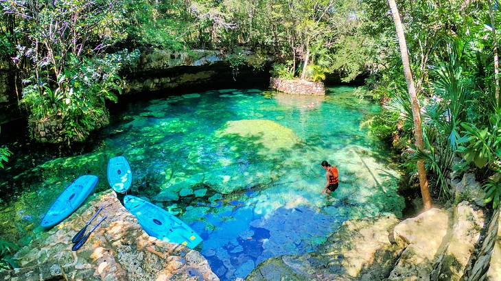 A person with a life vest in cenote water near blue kayaks and many green trees
