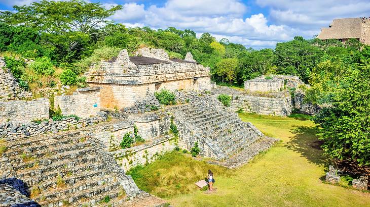 An aerial of a female standing in front of ruins with stairs near trees and grass